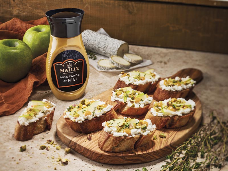 Moutarde au miel Maille présentée avec des tartines gourmandes au fromage de chèvre, pommes et pistaches, dans une ambiance chaleureuse inspirée de la cuisine française.
