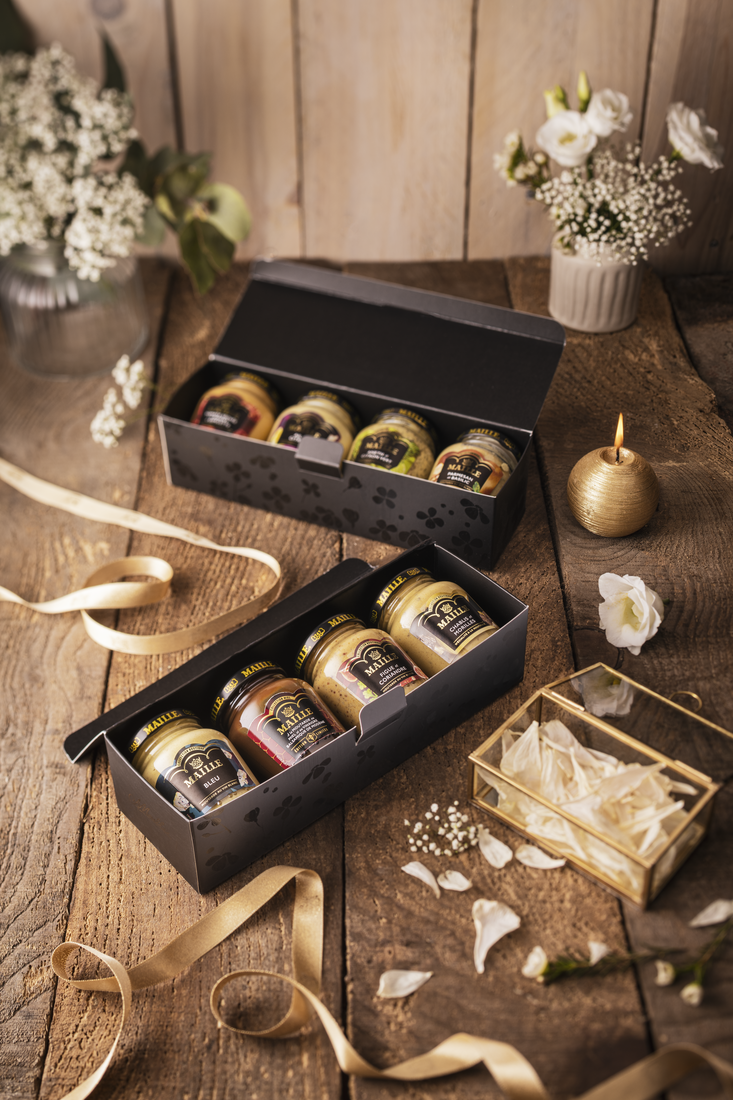 Deux coffrets cadeaux Maille ouverts, présentés sur une table en bois dans une ambiance chaleureuse et raffinée. Chaque coffret noir contient une sélection de moutardes Maille en pots de verre aux saveurs variées. La mise en scène est sublimée par des rubans satinés dorés, des fleurs délicates, des pétales dispersés et une bougie décorative, évoquant un univers élégant et gourmand idéal pour offrir.