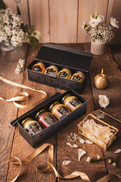 Deux coffrets cadeaux Maille ouverts, présentés sur une table en bois dans une ambiance chaleureuse et raffinée. Chaque coffret noir contient une sélection de moutardes Maille en pots de verre aux saveurs variées. La mise en scène est sublimée par des rubans satinés dorés, des fleurs délicates, des pétales dispersés et une bougie décorative, évoquant un univers élégant et gourmand idéal pour offrir.