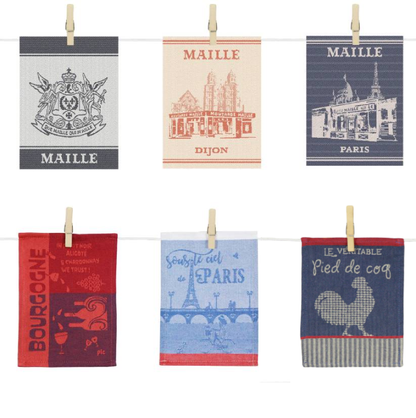 Collection de six torchons de cuisine suspendus à des cordes à linge à l’aide de pinces en bois. Chaque modèle présente un design distinct inspiré de l’univers Maille et du patrimoine français : blason Maille, illustration de la boutique historique de Dijon, boutique Maille Paris, motifs autour du vin et de la Bourgogne, vue stylisée de la tour Eiffel avec l’inscription « Sous le ciel de Paris » et illustration d’un coq accompagnée du texte « Le véritable pied de coq ». Les torchons arborent des teintes var