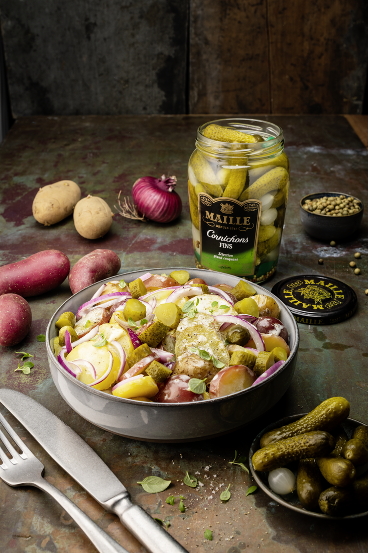 Cornichons fins Maille présentés dans un bocal en verre ouvert sur une table au style rustique. Le produit est mis en scène à côté d’une salade de pommes de terre garnie d’oignons rouges, de cornichons et d’herbes fraîches. Des pommes de terre, un oignon rouge, un bol de grains de poivre ainsi qu’une coupelle de cornichons complètent la composition. L’ambiance chaleureuse et authentique met en valeur le caractère traditionnel et gourmand des cornichons Maille.