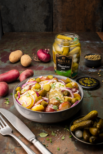 Cornichons fins Maille présentés dans un bocal en verre ouvert sur une table au style rustique. Le produit est mis en scène à côté d’une salade de pommes de terre garnie d’oignons rouges, de cornichons et d’herbes fraîches. Des pommes de terre, un oignon rouge, un bol de grains de poivre ainsi qu’une coupelle de cornichons complètent la composition. L’ambiance chaleureuse et authentique met en valeur le caractère traditionnel et gourmand des cornichons Maille.