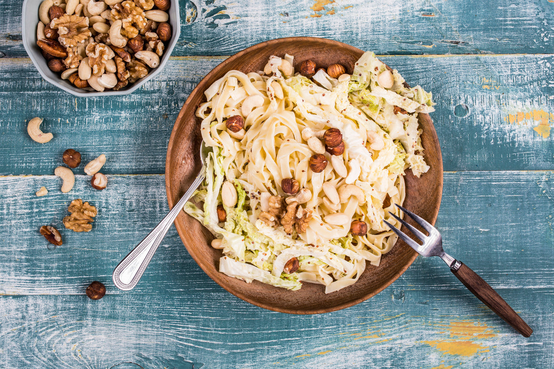 Salade de pâtes et chou chinois servie dans un grand bol en bois, garnie d'un mélange de fruits à coque comprenant noisettes, noix de cajou, amandes et noix. Le plat est présenté sur une table en bois bleu vieilli avec une fourchette et une cuillère. Un bol rempli de fruits secs est disposé à côté, tandis que quelques fruits à coque sont éparpillés autour de l'assiette pour une ambiance rustique et gourmande.