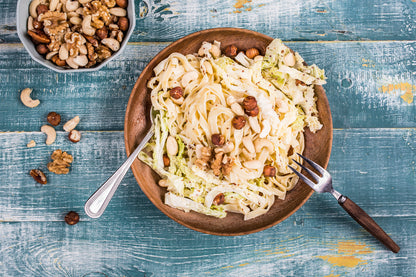 Salade de pâtes et chou chinois servie dans un grand bol en bois, garnie d'un mélange de fruits à coque comprenant noisettes, noix de cajou, amandes et noix. Le plat est présenté sur une table en bois bleu vieilli avec une fourchette et une cuillère. Un bol rempli de fruits secs est disposé à côté, tandis que quelques fruits à coque sont éparpillés autour de l'assiette pour une ambiance rustique et gourmande.