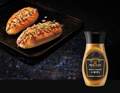 Deux sandwiches briochés garnis de viande effilochée, d’oignons rouges et d’herbes fraîches, nappés de moutarde au miel Maille. Les sandwichs sont présentés sur une plaque de cuisson noire dans une ambiance gourmande aux tons sombres. À droite du visuel, un flacon de moutarde au miel Maille est mis en avant, soulignant l’utilisation du produit dans la recette.