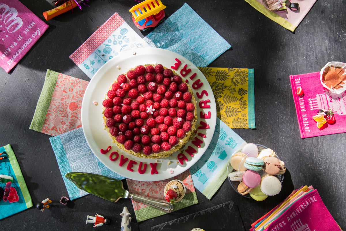 Gâteau d’anniversaire rond recouvert de framboises fraîches, présenté sur un plat blanc. L’inscription « Joyeux Anniversaire » est décorée en lettres rouges autour du gâteau. La table sombre est agrémentée de macarons colorés, d’une pelle à gâteau, de petites figurines décoratives et de serviettes aux motifs colorés. La composition festive et colorée met en scène un moment de célébration gourmand autour d’un dessert aux fruits rouges.