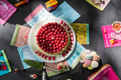 Gâteau d’anniversaire rond recouvert de framboises fraîches, présenté sur un plat blanc. L’inscription « Joyeux Anniversaire » est décorée en lettres rouges autour du gâteau. La table sombre est agrémentée de macarons colorés, d’une pelle à gâteau, de petites figurines décoratives et de serviettes aux motifs colorés. La composition festive et colorée met en scène un moment de célébration gourmand autour d’un dessert aux fruits rouges.