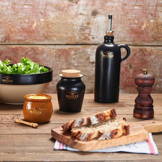 Photo lifestyle d'un pot à sauce miel, spatule 90mm, cruchon noir, saladier noir avec un poivrier et une planche à découper avec en dessous un para tapas 