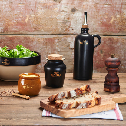 Photo lifestyle d'un pot à sauce miel, spatule 90mm, cruchon noir, saladier noir avec un poivrier et une planche à découper avec en dessous un para tapas 