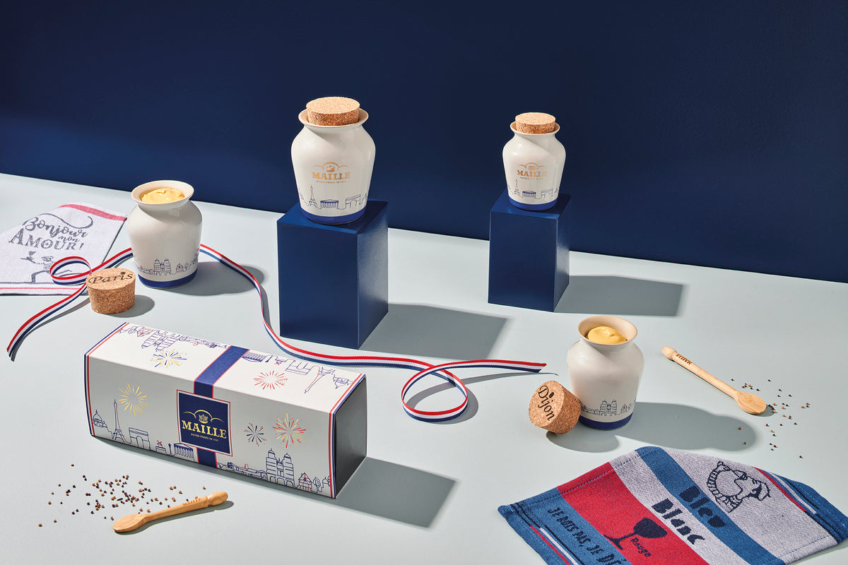 Coffret cadeau Maille avec pots en céramique blanche réutilisables, présenté dans une mise en scène aux couleurs bleu, blanc, rouge avec para-tapas et accessoires de dégustation.
