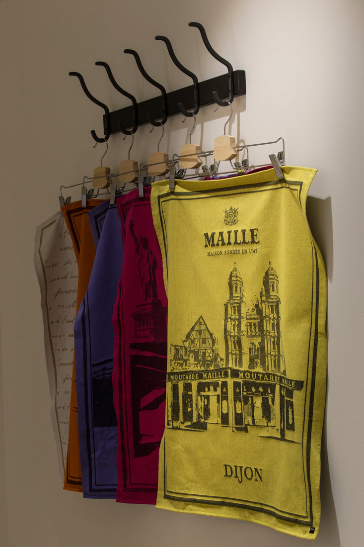 Torchons de cuisine décoratifs suspendus à un porte-manteau mural noir à plusieurs crochets. Au premier plan, un torchon jaune Maille illustré de monuments emblématiques de Dijon et de la boutique historique Maille, avec l’inscription « Dijon ». Derrière, plusieurs torchons colorés aux teintes orange, bleu, violet et rose sont suspendus sur des cintres. La scène est photographiée en intérieur sur fond clair, mettant en valeur la collection de torchons.