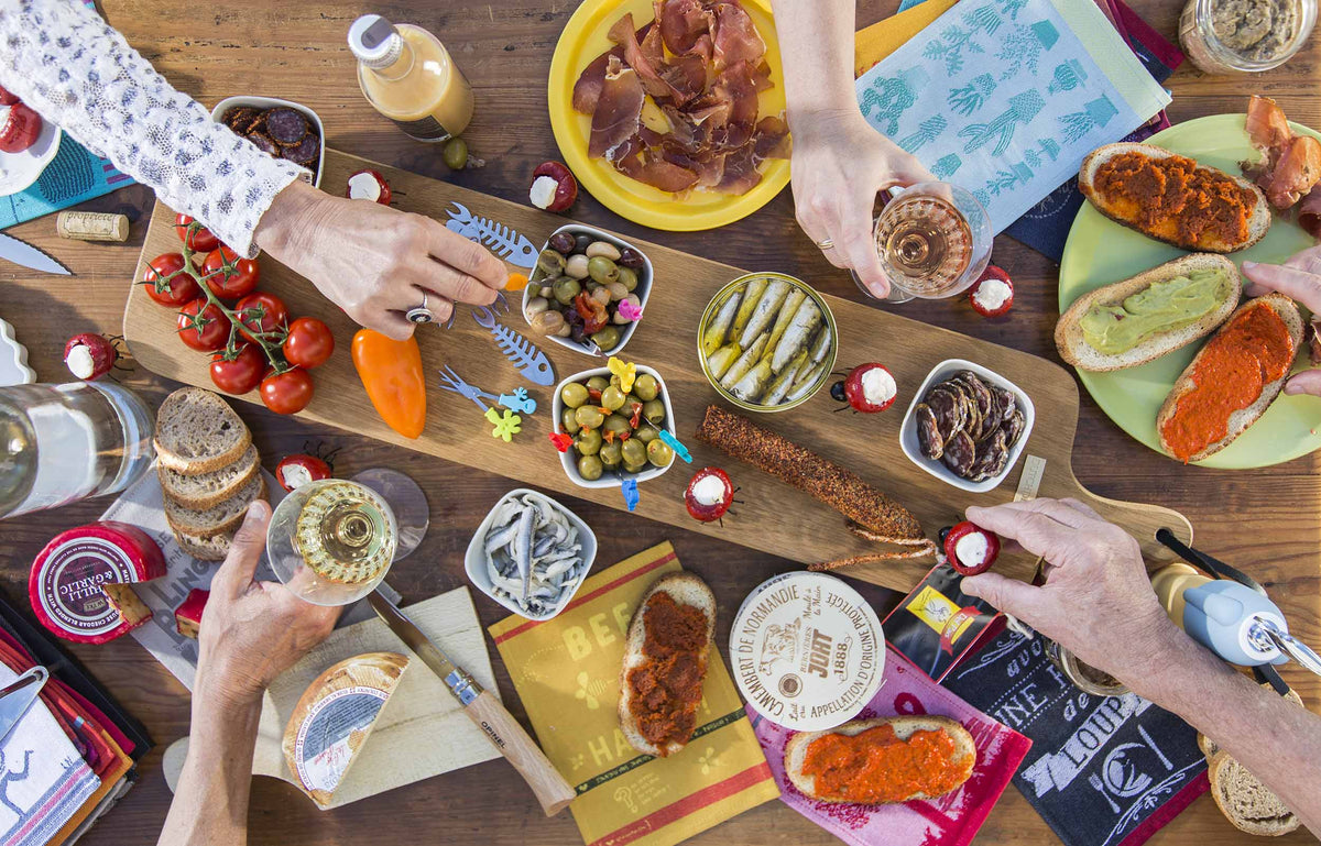 Table conviviale garnie de spécialités apéritives françaises, vue du dessus. Au centre, une planche en bois présente olives, sardines, saucisson, tomates cerises et légumes frais. Autour, plusieurs convives partagent des verres de vin, du fromage, du pain et des tartines gourmandes. Des torchons colorés et accessoires de table complètent cette scène chaleureuse inspirée des moments de partage et de la gastronomie française.