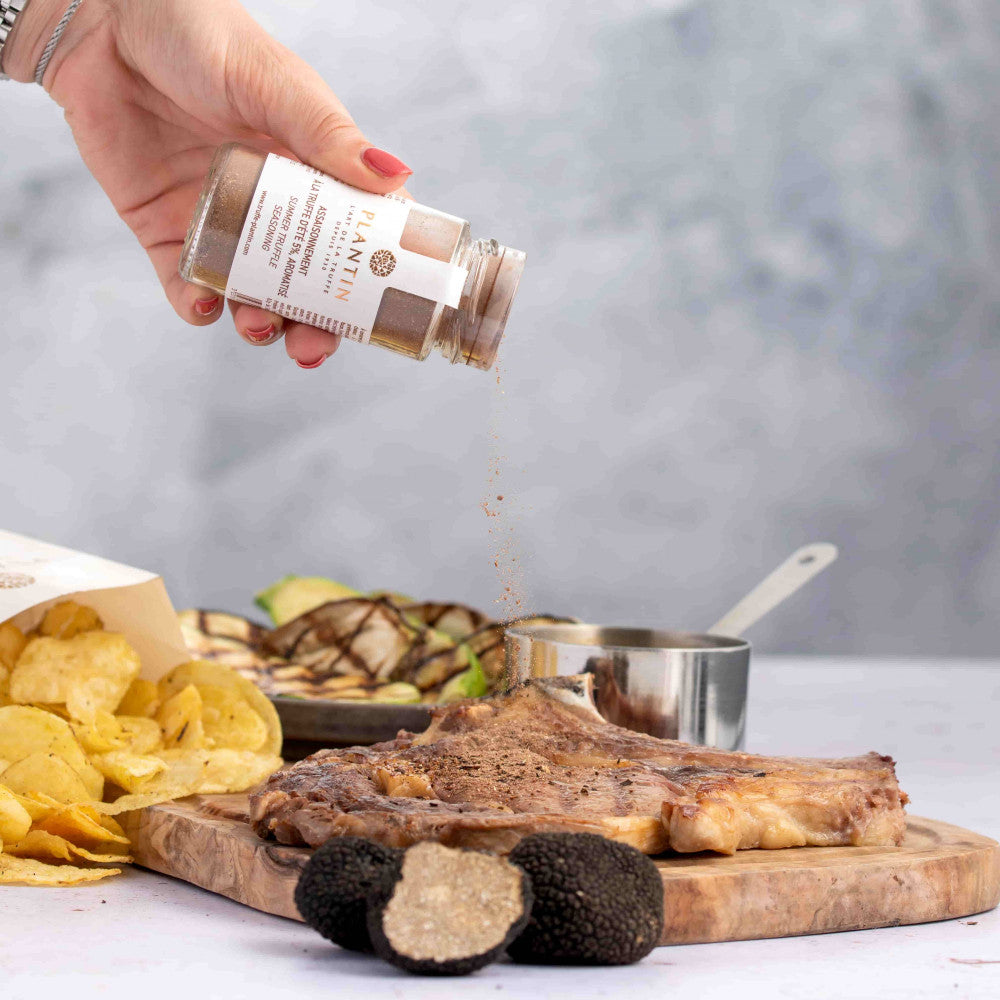 Assaisonnement à la truffe d’été Plantin saupoudré sur une pièce de viande grillée. Le flacon est tenu au-dessus du plat tandis que l’assaisonnement tombe sur la viande. La scène est complétée par des truffes fraîches, des chips artisanales et des légumes grillés dans une ambiance gastronomique et conviviale.