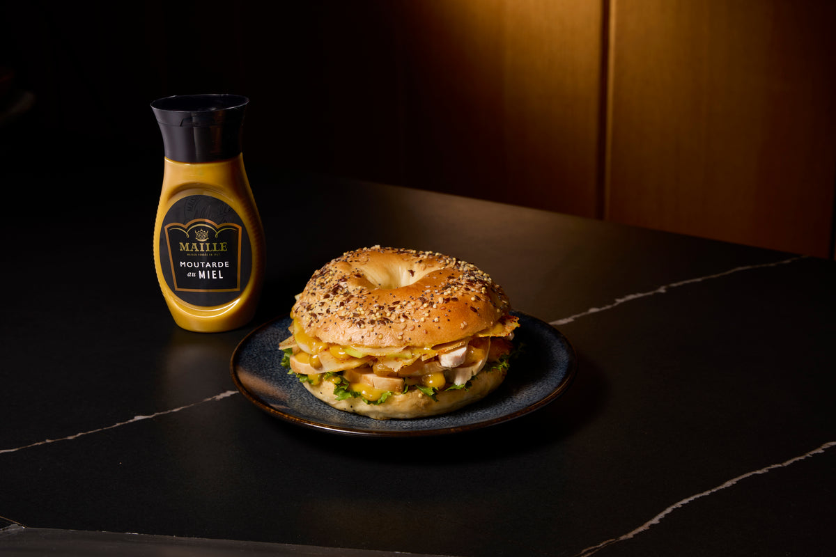 Moutarde au miel Maille présentée avec un bagel gourmand garni de poulet, fromage et légumes frais, dans une ambiance chaleureuse et raffinée.