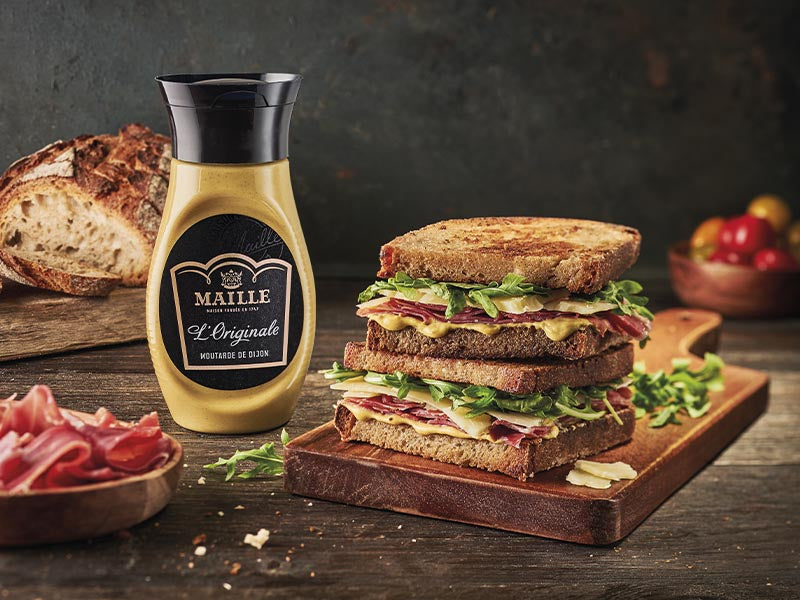 Moutarde de Dijon Maille L'Originale présentée avec des clubs sandwichs gourmands garnis de jambon, fromage et roquette, dans une ambiance chaleureuse inspirée de la gastronomie française.