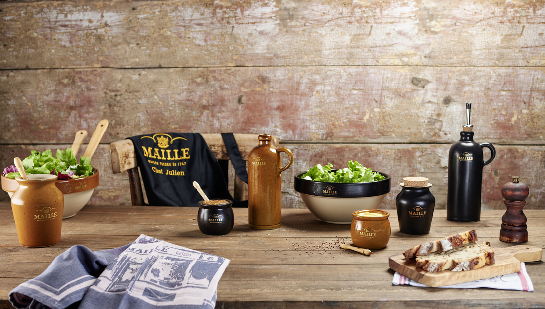 Collection Art de la Table Maille présentée sur une grande table en bois rustique dans une ambiance chaleureuse et authentique. La mise en scène rassemble plusieurs pièces en grès, dont des saladiers garnis de salade, des cruches, des moutardiers, un huilier noir, un pot à sel avec couvercle en liège, un moulin à poivre et du linge de table Maille. Une planche de pain de campagne complète cet univers inspiré de l’art de recevoir à la française.