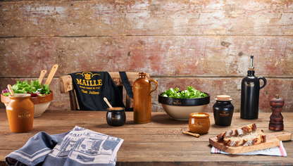 Collection Art de la Table Maille présentée sur une grande table en bois rustique dans une ambiance chaleureuse et authentique. La mise en scène rassemble plusieurs pièces en grès, dont des saladiers garnis de salade, des cruches, des moutardiers, un huilier noir, un pot à sel avec couvercle en liège, un moulin à poivre et du linge de table Maille. Une planche de pain de campagne complète cet univers inspiré de l’art de recevoir à la française.