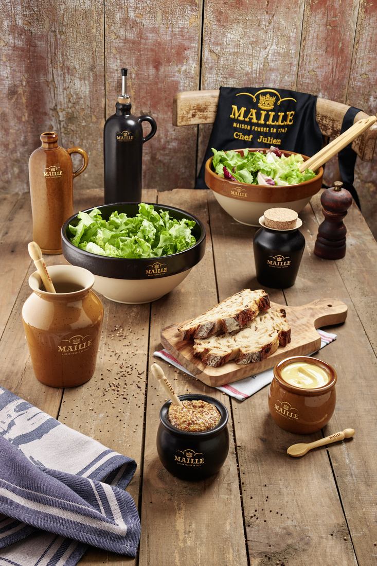 Collection d’art de la table Maille en grès de Digoin présentée sur une table en bois rustique. L’ensemble comprend des saladiers garnis de salade verte, des moutardiers, des huiliers, un moulin à poivre, des cuillères de service et une planche de pain de campagne. Un tablier Maille noir brodé complète cette mise en scène chaleureuse inspirée de l’art de recevoir à la française.