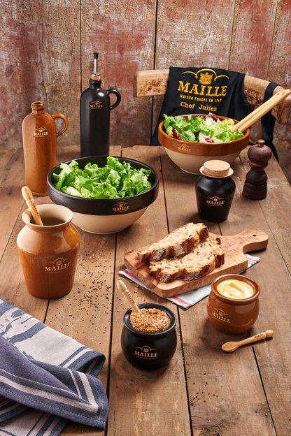 Collection d’art de la table Maille en grès de Digoin présentée sur une table en bois rustique. L’ensemble comprend des saladiers garnis de salade verte, des moutardiers, des huiliers, un moulin à poivre, des cuillères de service et une planche de pain de campagne. Un tablier Maille noir brodé complète cette mise en scène chaleureuse inspirée de l’art de recevoir à la française.