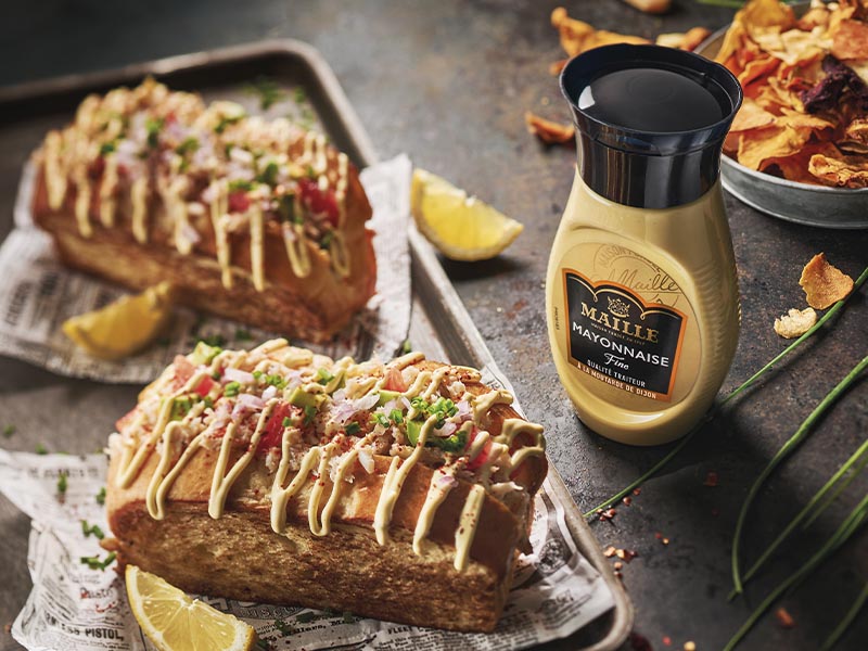 Mayonnaise Fine Maille à la moutarde de Dijon présentée avec des hot-dogs gourmands garnis de légumes frais et d'herbes aromatiques, dans une ambiance conviviale et raffinée.