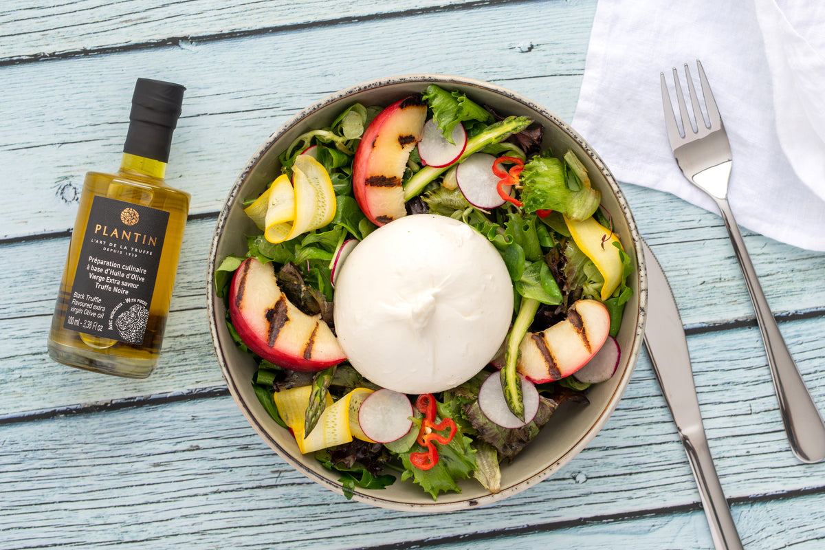Bouteille d’huile d’olive vierge extra Plantin aromatisée à la truffe noire présentée aux côtés d’une salade gourmande composée de burrata, jeunes pousses, radis, asperges, légumes frais et fruits grillés. La scène est disposée sur une table en bois clair dans une ambiance fraîche et raffinée.
