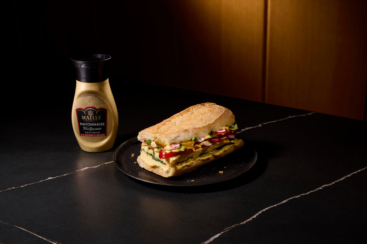 Mayonnaise Maille Fins Gourmets à la moutarde à l’ancienne présentée avec un sandwich artisanal garni de légumes frais, dans une ambiance gourmande et raffinée.