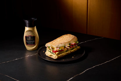 Mayonnaise Maille Fins Gourmets à la moutarde à l’ancienne présentée avec un sandwich artisanal garni de légumes frais, dans une ambiance gourmande et raffinée.