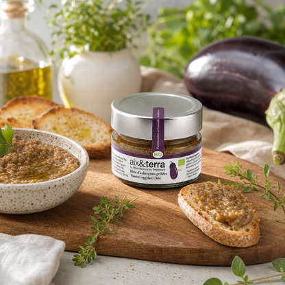 Pot de riste d’aubergines grillées bio Aix & Terra présenté sur une planche en bois dans une ambiance provençale chaleureuse. Le produit est entouré de tartines garnies de préparation aux aubergines, d’aubergines fraîches, d’huile d’olive et d’herbes aromatiques. Un bol de riste d’aubergines complète cette mise en scène gourmande inspirée des saveurs méditerranéennes.