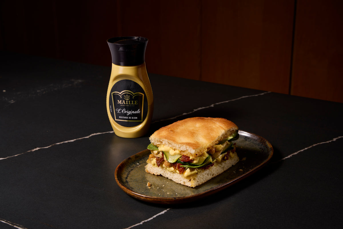 Moutarde de Dijon Maille L'Originale présentée avec un sandwich gourmand garni de légumes frais et de fromage, dans une ambiance chaleureuse et raffinée.