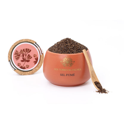 Pot de sel fumé Les Terres Blanches ouvert, révélant des cristaux de sel foncés. Le contenant en céramique couleur terracotta est accompagné de son couvercle en liège décoré d’un motif floral et d’une cuillère en bois remplie de sel fumé.