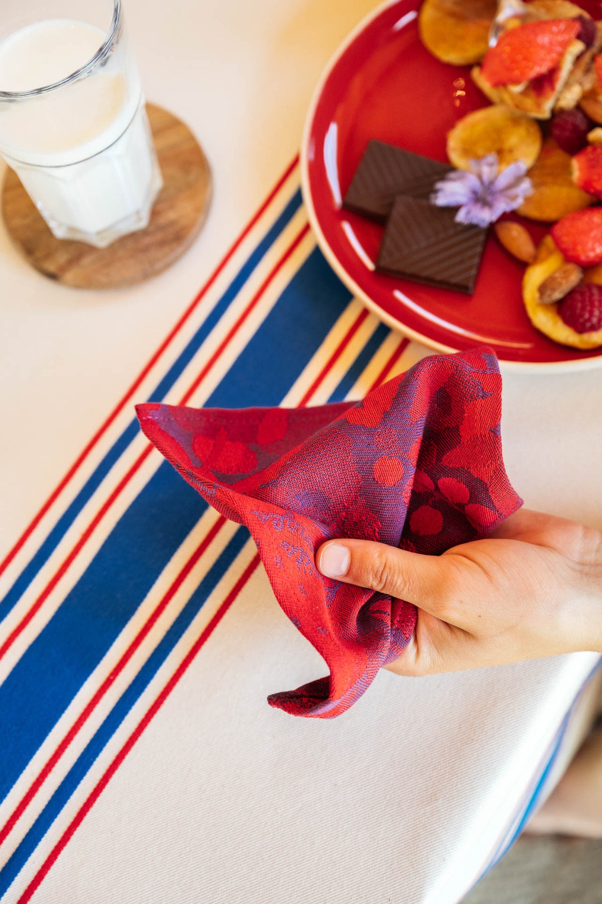 Main tenant une serviette de table Maille aux tons rouge et bleu, ornée d’un motif tissé à pois et détails floraux. La serviette est présentée sur une nappe blanche à rayures bleu, blanc et rouge. À proximité, une assiette rouge garnie de pancakes, fraises fraîches, fruits secs et carrés de chocolat noir, ainsi qu’un verre de lait posé sur un sous-verre en bois, complètent cette scène de petit-déjeuner gourmande et conviviale.