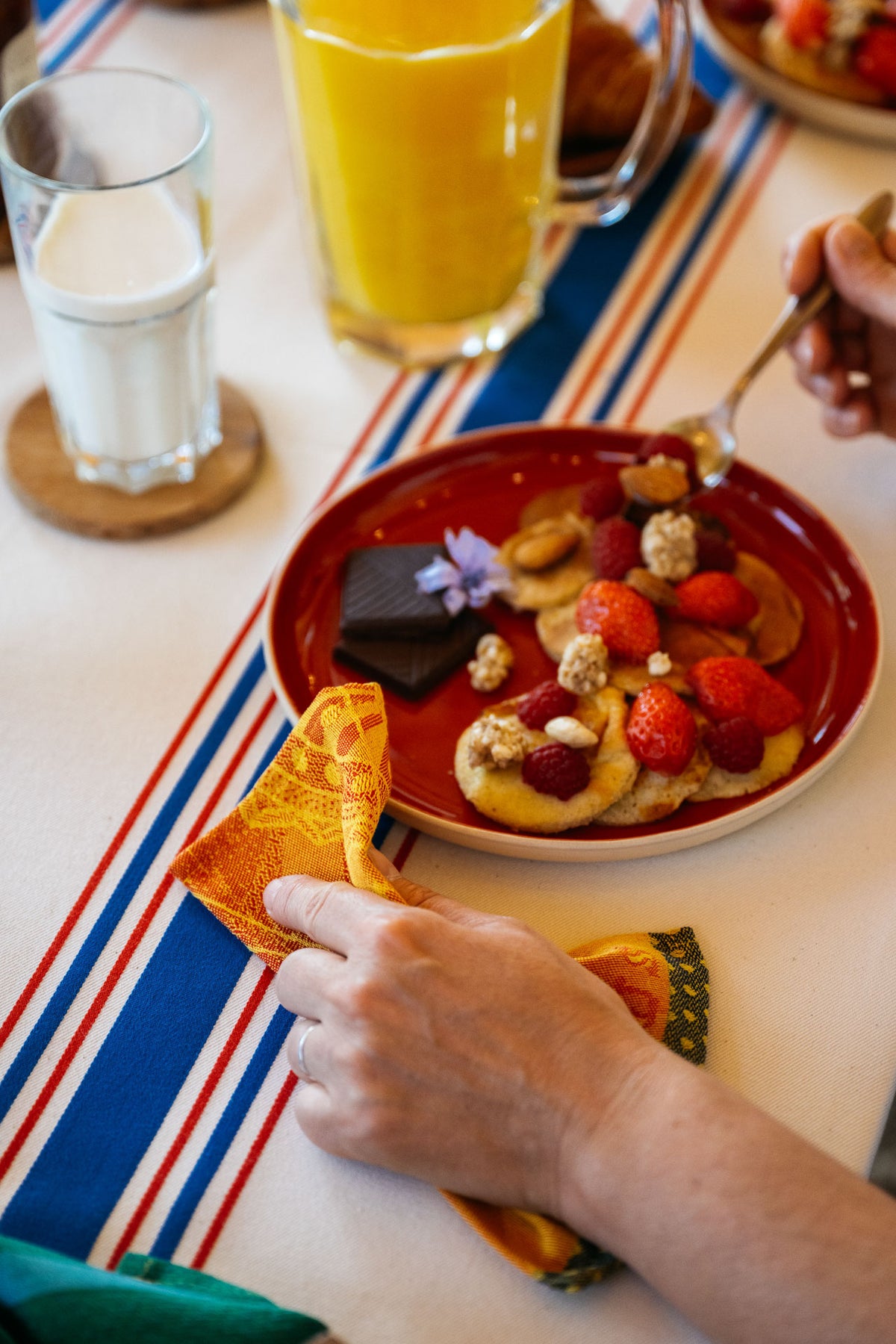 Table de petit-déjeuner gourmande avec une assiette rouge garnie de pancakes, fraises fraîches, framboises, fruits secs, céréales croustillantes et carrés de chocolat noir. Au premier plan, une main tient une serviette de table Maille aux tons orange et jaune à motif tissé. Une autre main utilise une cuillère au-dessus de l’assiette. Un verre de lait et une carafe de jus d’orange sont disposés sur une nappe blanche ornée de rayures bleu, blanc et rouge, créant une ambiance conviviale et colorée.
